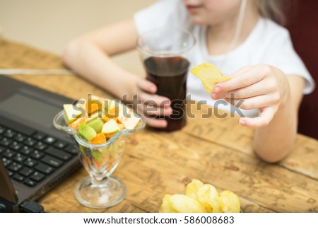 Girl eats potato chips and fruit salad in front of laptop. Unhealthy eating in front of computer.