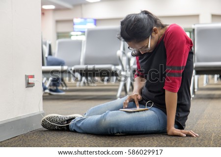 A young woman sit on floor and recharges the phone from the wall socket in the hall. Passengers relaxed in the waiting room at the airport. Girl with mobile phone in the lobby.