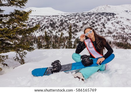 Beautiful snowboard girl with a crown on the head standing on the top of the mountain holding snowboard. Queen of snowboard. Winner of a beauty contest.