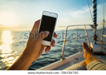 Summer leisure. Rest on a yacht with a phone in his hand. Man lying on the deck and enjoy your smartphone. The guy doing the photo feet on the background seascape and yachts. Royalty-Free Stock Photo #586186760