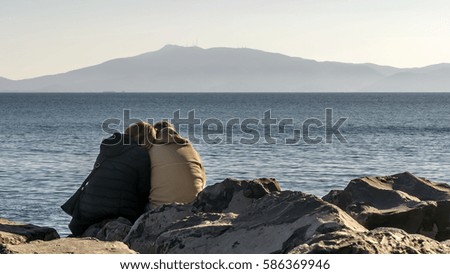 A loving couple cuddles on the rocks in front of the sea in winter and with the coast in the background, Talamone, Grosseto, Tuscany, Italy