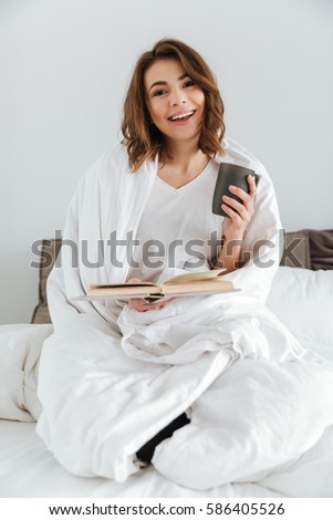 Picture of young pretty lady dressed in white t-shirt sitting on bed at home indoors while reading book and drinking coffee.