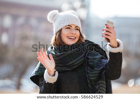 Image of attractive young lady walking on the street wearing hat and scarf while using mobile phone and waving.