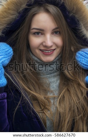 Beautiful girl with long hair looking into the camera in a warm jacket. She is dressed in a purple jacket and blue mittens.