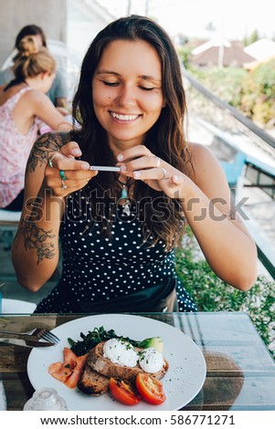 Young woman taking picture of her breakfast made of poached eggs benedict served on toast with smoked salmon, avocado, grilled tomato and spinach, with smart phone 