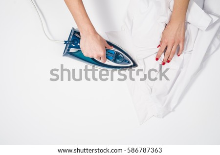 Female hand ironing clothes top view isolated on white background. Young woman with iron ironing man's shirt seen from above during housework. Blue iron on white table. Royalty-Free Stock Photo #586787363
