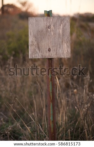 Blank unlettered weathered plywood sign on rusting and chipped paint ranch pole in shade of dusk with shallow depth of field in front of golden tall grass and California chaparral shrub field
