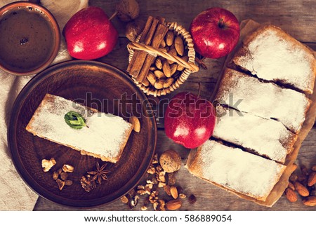 Sliced homemade apple strudel served with fresh apples, cinnamon sticks and sugar powder over old wooden background. Close up, dark rustic style Apple strudel.
