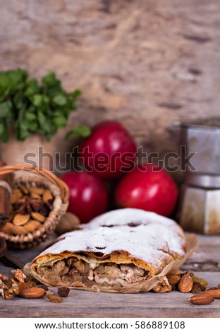 Sliced homemade apple strudel served with fresh apples, cinnamon sticks and sugar powder over old wooden background. Close up, dark rustic style Apple strudel.