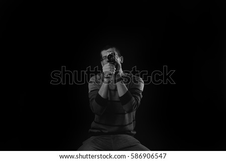 Young man standing on a chair and looking true an old camera on dark background. Black and white