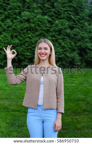 Slender blonde smiling and shows gesture "OK", enjoying life and new day, advertises any product shows emotion and gesture. Women with long blonde hair dressed in bright blouse, beige jacket and blue