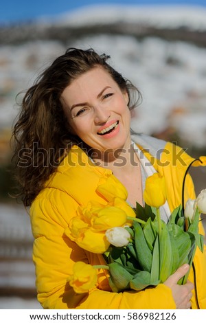 Charming woman with a bouquet of tulips against the background of snow-covered mountain tops. She is dressed in a bright yellow down-padded coat. Woman has a happy look.
