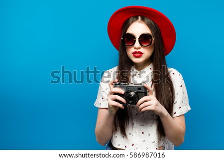 Fashion pretty young smiling woman holds retro camera wearing red hat, over blue background