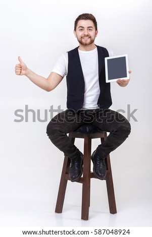 young handsome man with beard in white shirt and black waistcoat holding tablet and showing thumb up sign on gray background