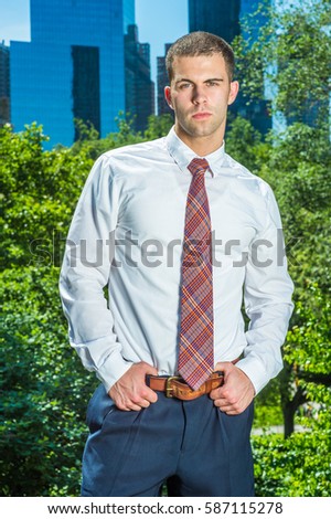 Portrait of Businessman in New York. Handsome professional guy wearing long sleeve white shirt, patterned tie, hands on belt, stands in font of business district, ready to work.
