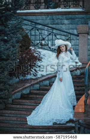 Beautiful lady in vintage white dress and a hat with feathers near her estate. Edwardian period wedding dress. 