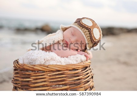 Two week old newborn baby boy sleeping in a wicker basket wearing an aviator hat. Photographed on a beach with a rock jetty. Royalty-Free Stock Photo #587390984