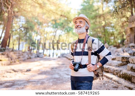 Enjoying travel. Senior smiling man with backpack holding camera on ancient sightseeing background.