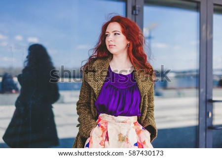 Cool cute redhead woman posing in a stylish green coat, purple t-shirt and bright beautiful lush skirt on the big city background