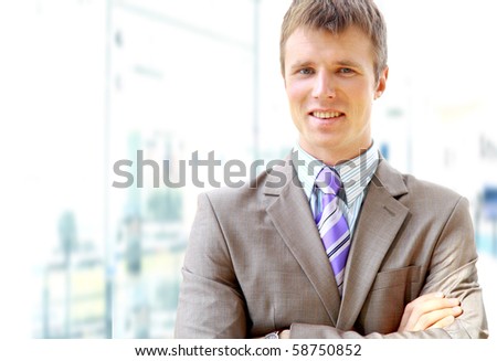 Happy businessman standing with hands in pocket in front of windows, looking at camera, smiling