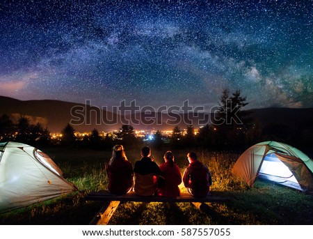 Silhouette of four people sitting on a bench made of logs and watching fire together beside camp and tents in the night. On the background starry sky, Milky way, mountains and luminous town. Rear view