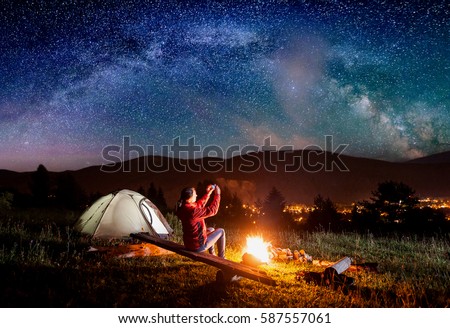 Hiker woman sitting on boards by a campfire near tent and makes the photo incredible view of night sky with stars and Milky way on the phone