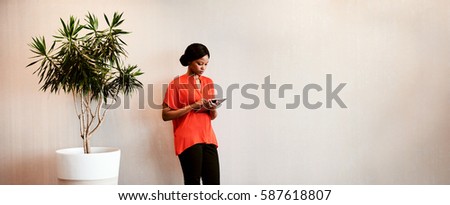 Black businesswoman holding a digital tablet in her hands while standing next to a tree planted in a pot against a textured wall with text space to the right of the image.