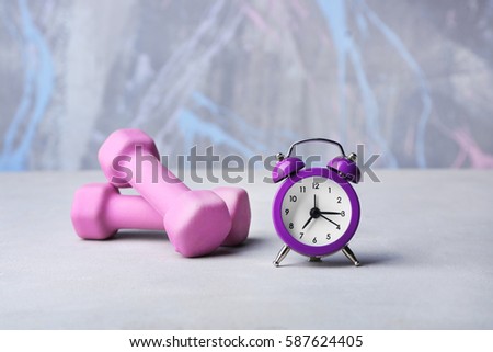 Alarm clock and dumbbells on color background