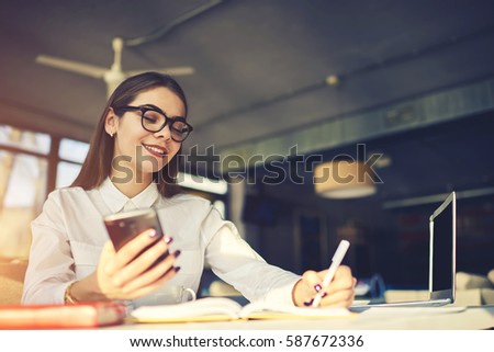 Cheerful young female freelancer felling happy to receive message from client suggesting working project noting his contact to make interview and discuss details of collaboration while using cafe wifi