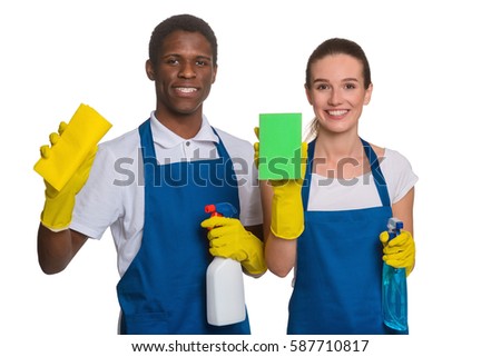 Black male holding a spray and a rag. White female in working clothes holding a sponge and a bottle. Two smiling janitors in a cleaning uniform standing by each other.
