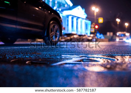 Rainy night in the big city, the car rides near the illuminated luxurious mansion. Close up view of a hatch at the level of the asphalt, image in the blue tones