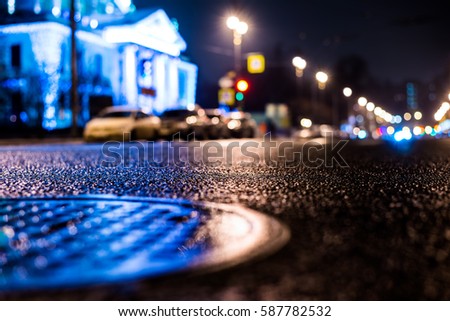 Rainy night in the big city, the road passes next to the illuminated luxurious mansion. Close up view of a hatch at the level of the asphalt, image in the blue tones