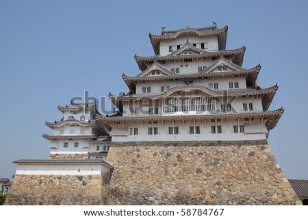 Landscape view of the main tower of Himeji Castle on the hillside during the daytime with bonsai pine trees of the castle gardens in the foreground and blue sky in the background