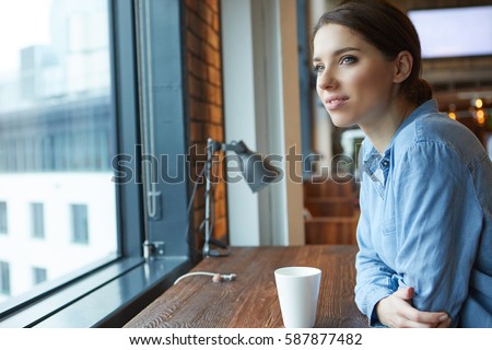 Young attractive girl  sitting alone near big window in coffee shop during free time and working 