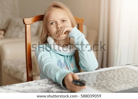 Portrait of adorable blonde female child in warm cozy clothing clicking remote control buttons, flipping through channels, looking for cartoons, having concentrated look, sitting in domestic interior