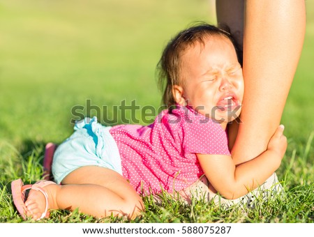 Crying baby girl holding onto mothers leg outdoors. Royalty-Free Stock Photo #588075287