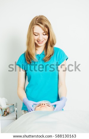Portrait of young blond woman cosmetologist in blue t-shirt doing face cleaning in European spa salon on white background. Facial massage for young woman. Photo series with real people models.