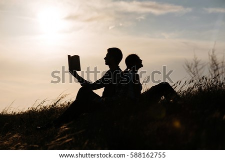 man with girl sitting on grass and read a book. silhouette of couple at sunset