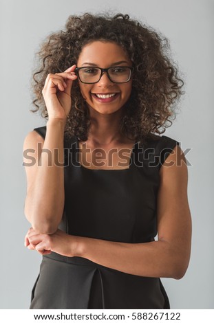 Beautiful Afro American girl in black cocktail dress and eyeglasses is looking at camera and smiling, on gray background