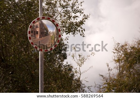 Convex round traffic mirror ringed with red and white checkers above on metal street lamp post reflecting brick building and rural street scene with deciduous tree leaves on branches and cloudy sky 