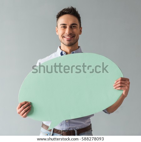 Attractive young man is holding a speech bubble, looking at it and smiling, on gray background