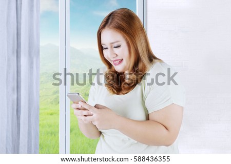 Picture of happy blonde woman with overweight body, texting on her smartphone while standing near the window