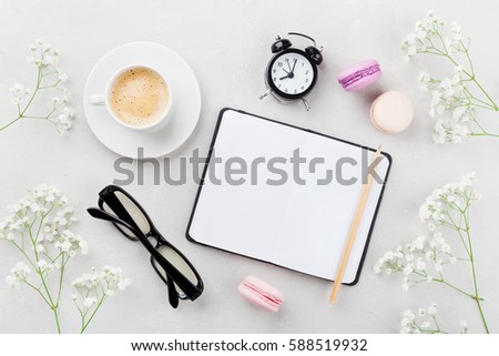 Coffee, cake macaron, clean notebook, eyeglasses, alarm clock and flower for cozy breakfast on light table top view. Woman working desk. Flat lay style