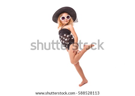 Isolated on white, little cute caucasian blonde girl in black swimwear, white sunglasses and big black hat stand, look at camera, one leg up
