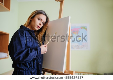 Attractive young woman artist begins to draw a new paint on a  wooden easel, holds brush.