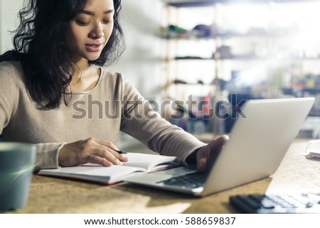 Portrait of an inspired Asian office employee looking at her notes and entering data into computer system.
