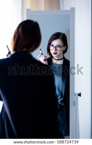 beautiful young woman standing in front of mirror, holding a lipstick and looking at her reflection