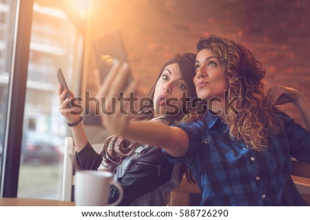 Two friends drinking coffee in a cafe, taking selfies with a smart phone and having fun making funny faces. Focus on the girl on the left