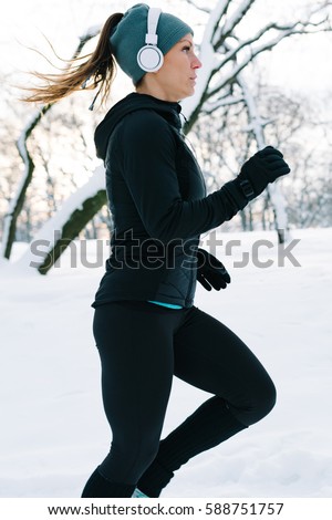 Female athlete jogging in park in winter with snow around the park. Listeninig music and exercising