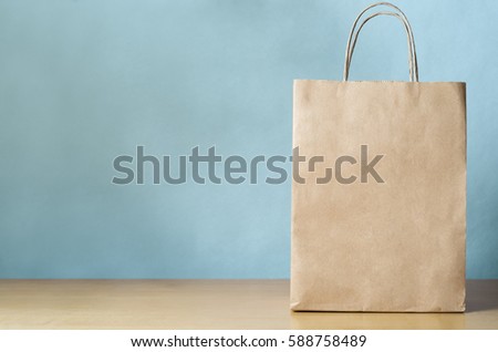 Blank brown paper carrier bag with handles for shopping, facing front on right side of a light wood veneer table with pale blue wall background providing copy space to left. Royalty-Free Stock Photo #588758489
